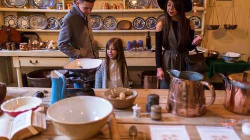 Three visitors stand around a table in a historic kitchen display, examining traditional cooking equipment. The room contains copper pots, ceramic mixing bowls, weighing scales, eggs and kitchen utensils on a wooden worktable, with shelves of blue-and-white plates and copper cookware lining the walls behind them. Warm indoor lighting highlights the period furnishings and collection of household objects.